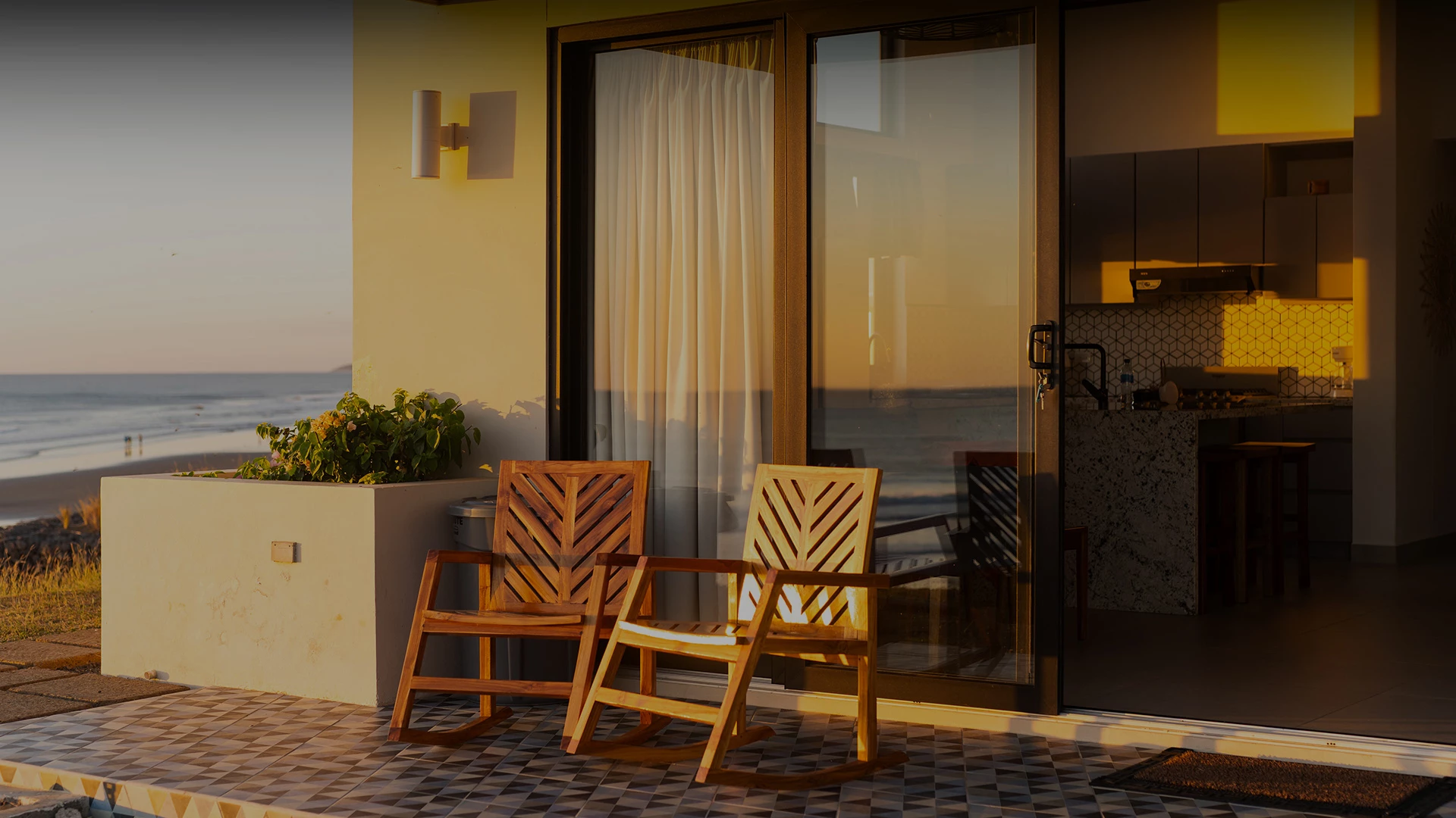 Two wooden chairs sit on a tiled patio by a glass door, with a beach/ocean view and a warm kitchen inside.