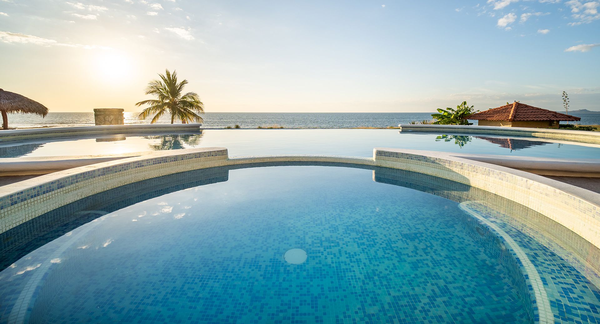 A beachfront villa with a blue tiled infinity pool overlooking the ocean at sunrise, palm trees and rooftops in view.