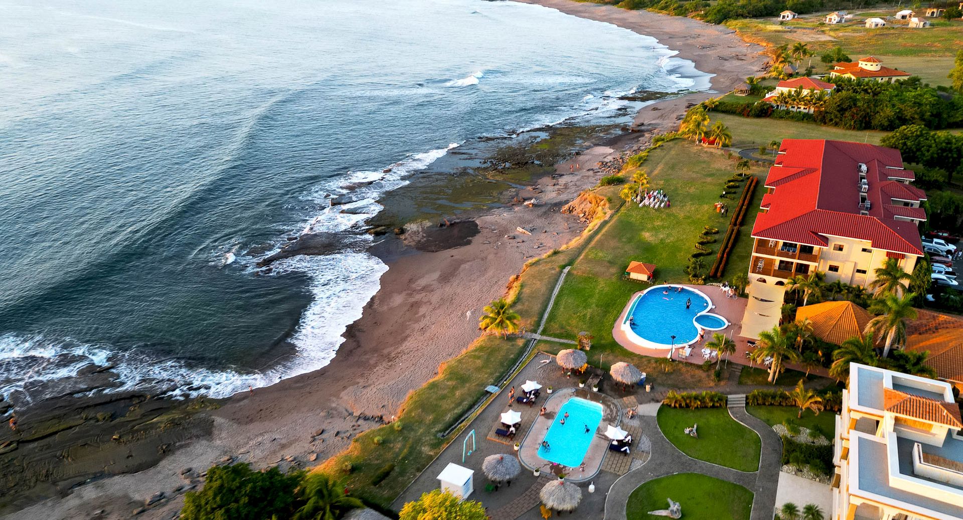Aerial view of a coastal resort with red-roof buildings, a sandy beach, ocean waves, and outdoor pools along a lush shoreline.