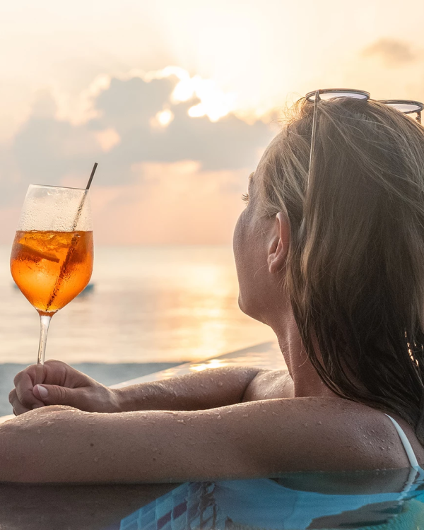A woman lounges on a surfboard at sunset, holding a bubbly orange cocktail in a stemmed glass.