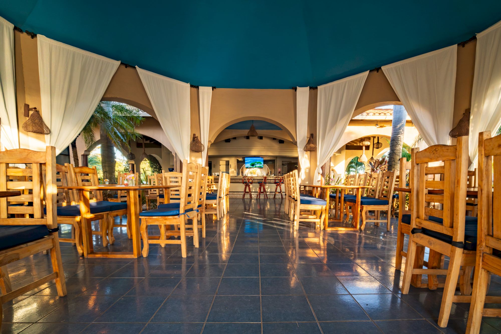 Sunlit restaurant pavilion with white curtains, wooden chairs, arches, pendant lights, and a TV&mdash;tropical courtyard feel.