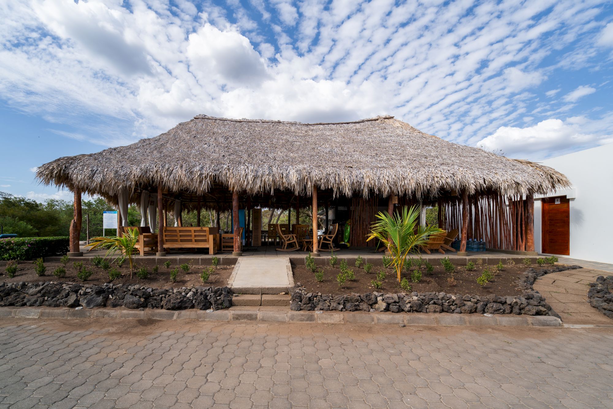A thatched outdoor pavilion with wooden benches and palm plants under a blue sky with scattered clouds.
