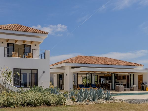A modern white two-story house with terracotta roof, patio dining, pool area, desert landscaping, under a clear blue sky.