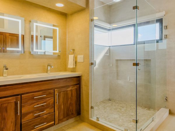 A modern bathroom with a wooden vanity, two illuminated mirrors, and a walk-in glass shower with beige tile flooring.