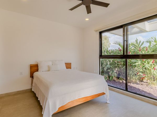 A modern bedroom with a wooden bed, white walls, tiled floor, ceiling fan, and large sliding glass doors opening to lush plants.