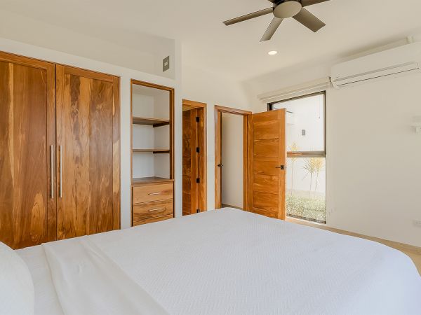 A bright bedroom with a white bed, wooden closet doors, open shelving, ceiling fan, and wall air conditioner above the window.