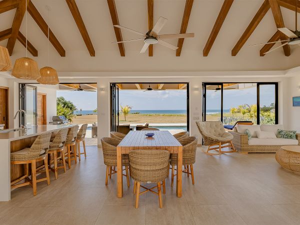 Bright beachside open-plan living and dining area with wicker furniture, ceiling fans, pendant lights, and an ocean view.