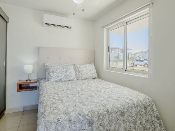 Bright bedroom with a double bed, grey patterned bedding, wall AC, bedside lamp, and a window showing parked cars.