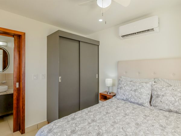 Modern bedroom with a patterned bed, ceiling fan, wall A/C, plus a visible bathroom sink and round mirror.