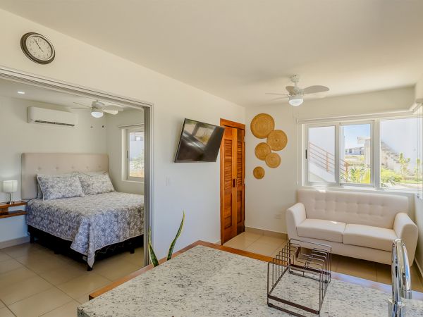 Bright modern living room with white sofa, dining table, beach wall art, big windows, and an adjacent bedroom with bed and clock.