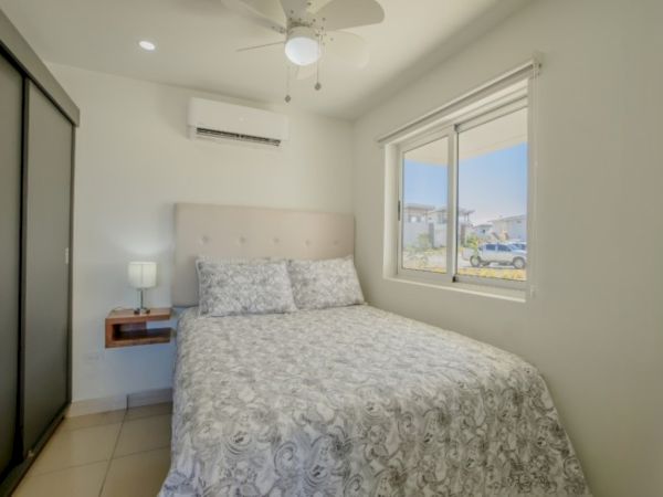 A bright bedroom with a bed, ceiling fan, bedside lamp, and large window looking out to a street.