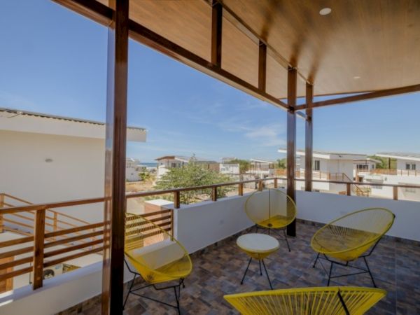 A sunny balcony patio with a wooden pergola, yellow chairs, and a view over a seaside neighborhood to the horizon.