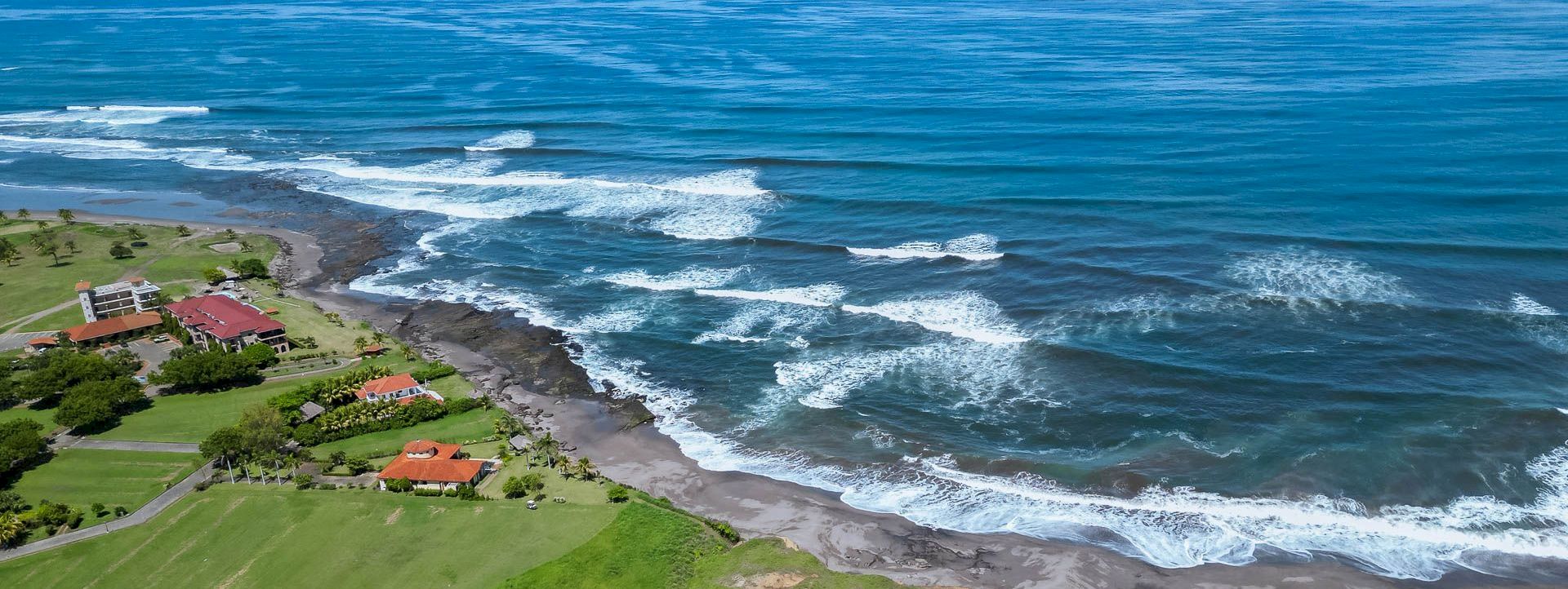Aerial view of a coastal resort with green lawns, a sandy beach, and waves crashing along the shoreline.