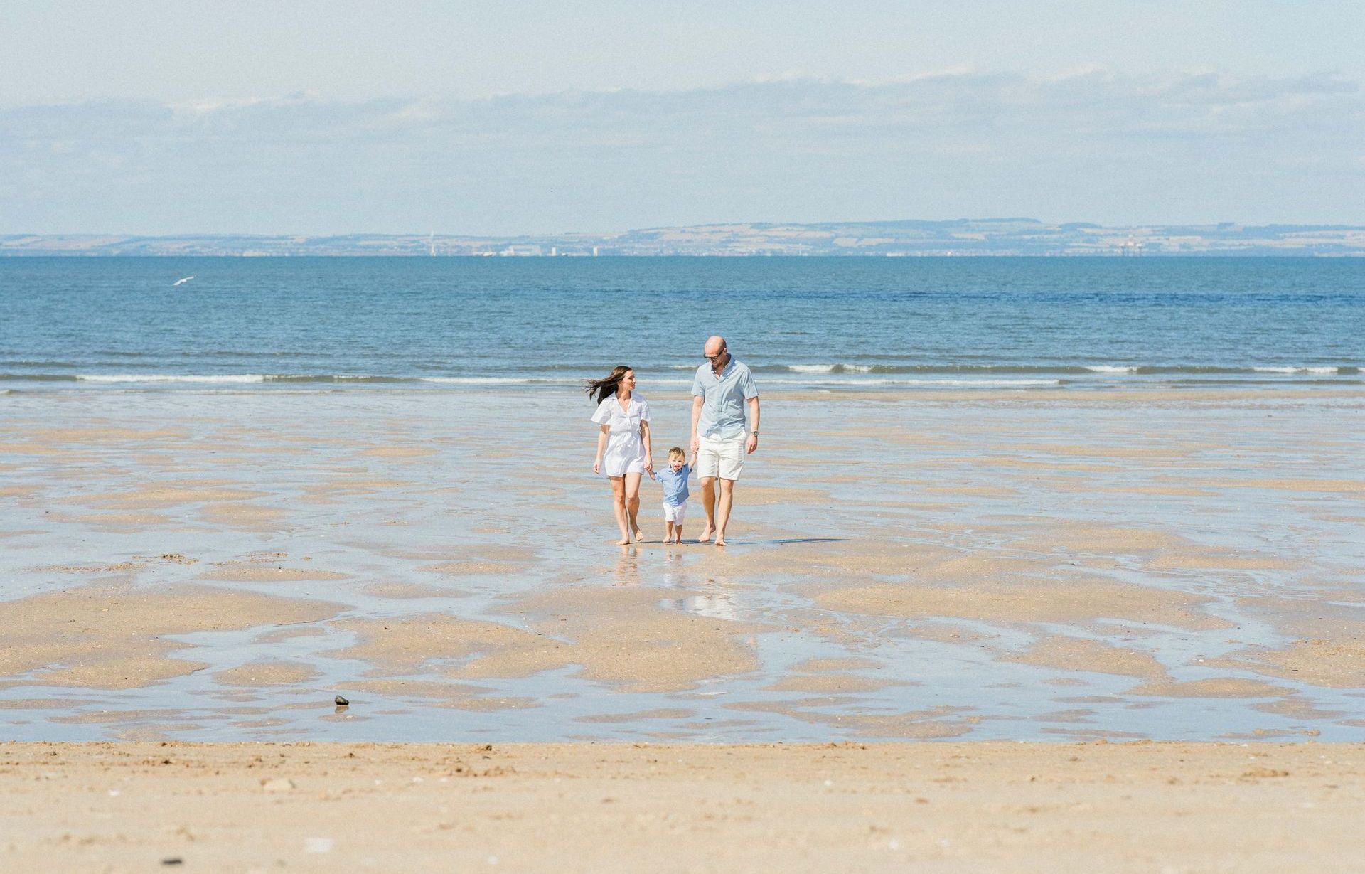 A family of three walks along a sunny shoreline, holding hands and strolling in the waves&rsquo; reach.