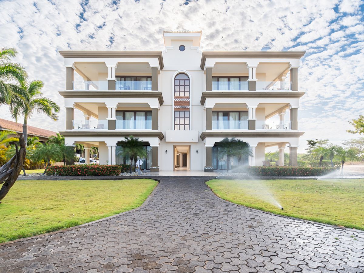 A grand white villa with balconies, arches, manicured lawns, and palm trees under a bright sky.