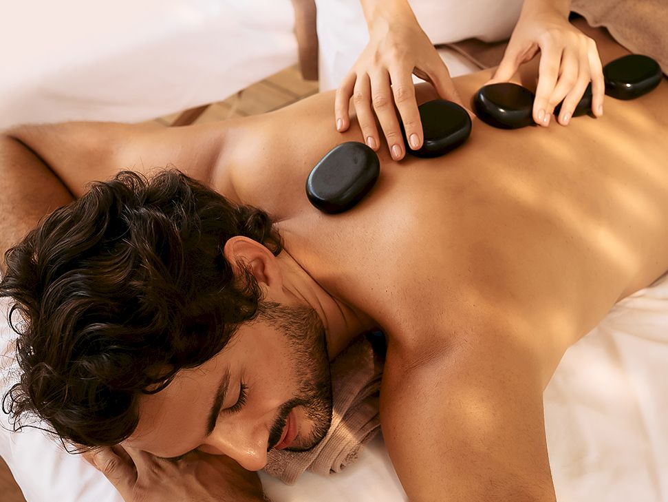 A spa massage: hot stones rest on a man&rsquo;s back while a therapist&rsquo;s hands gently glide over his skin.