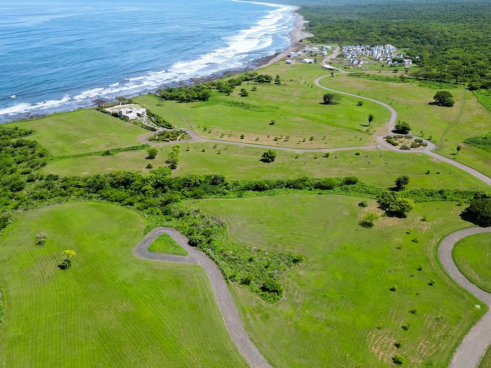 Aerial view of green coastal countryside with winding roads, scattered trees, and a small beachfront resort along the ocean.
