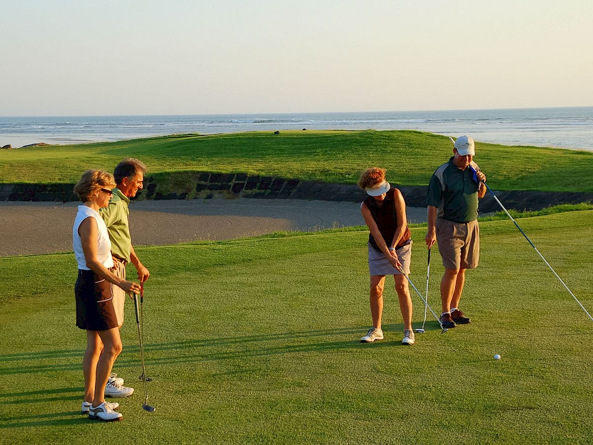 A group of golfers practices on a grassy course by the ocean, holding clubs and putting under sunny skies.