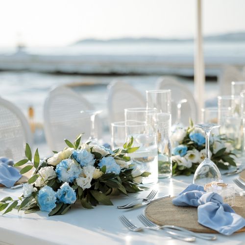 A seaside table set for an event: blue and white floral centerpiece, crystal glasses, blue napkins, and cutlery by the water.