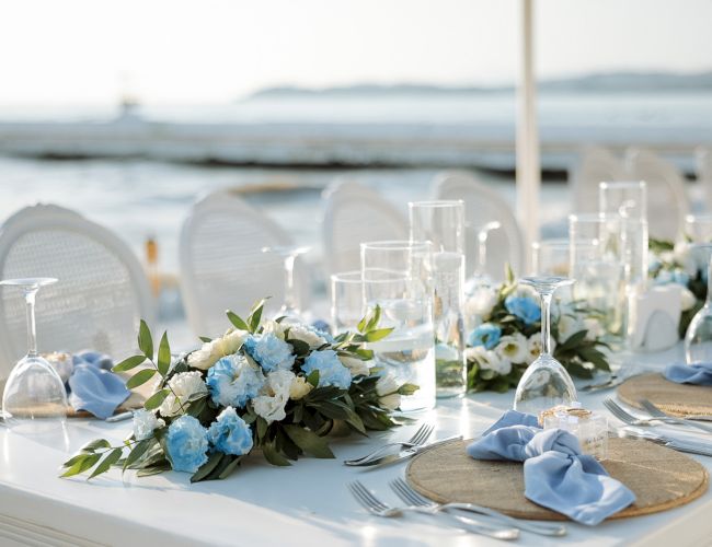 A seaside table set for an event: blue and white floral centerpiece, crystal glasses, blue napkins, and cutlery by the water.