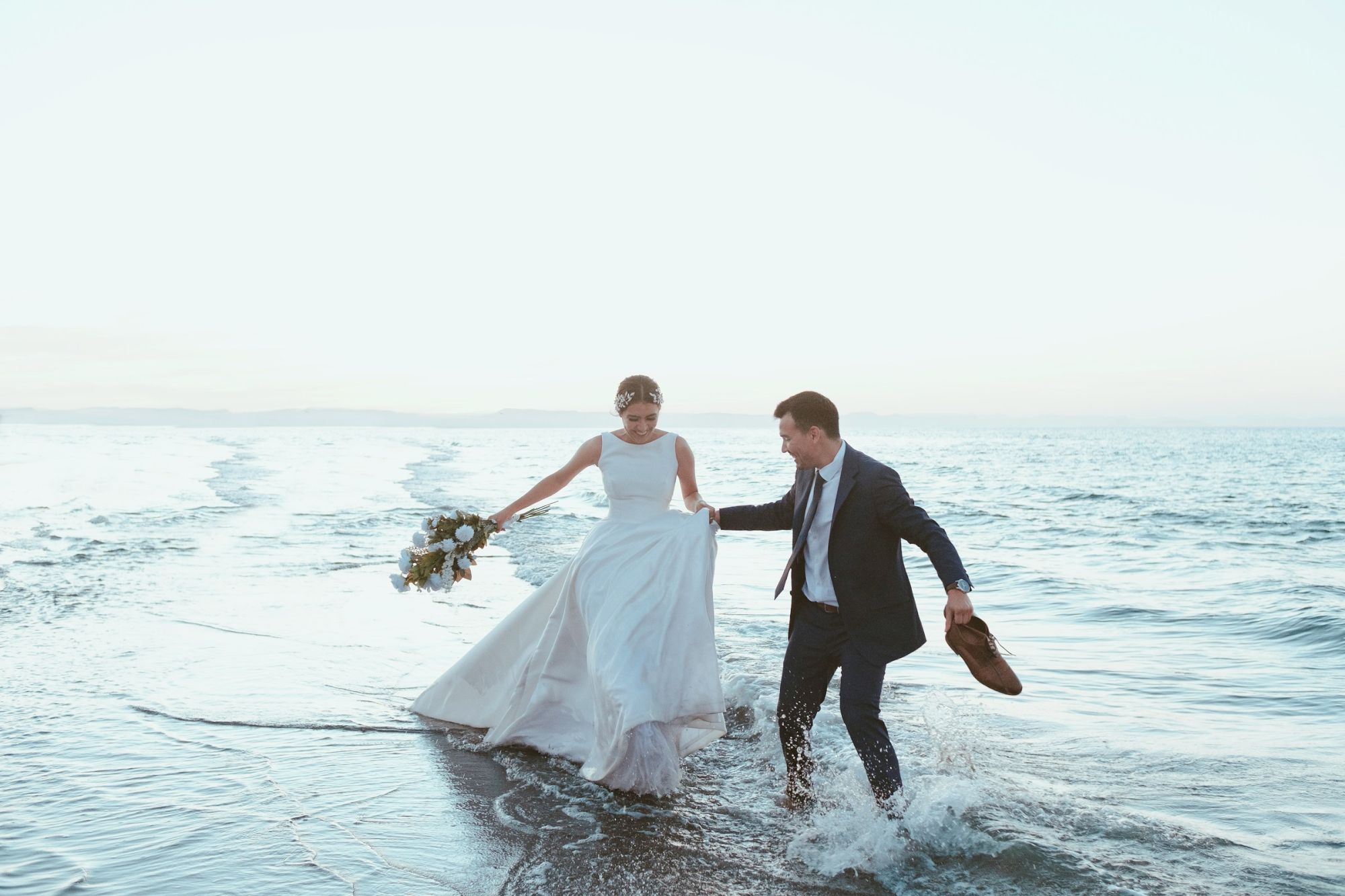 A bride in a white dress and a groom in a suit walk in ocean surf on a beach, holding her bouquet and his hand.