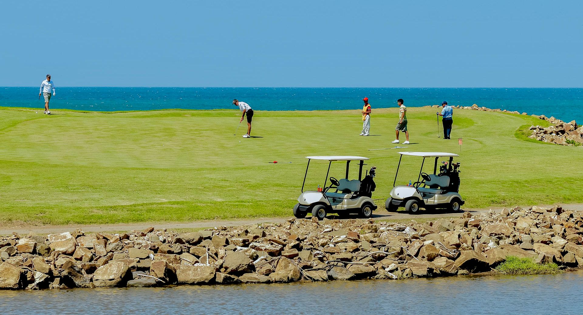 Golfers play on a sunny seaside course, with a couple of golf carts parked by a rocky shoreline and ocean in the background.