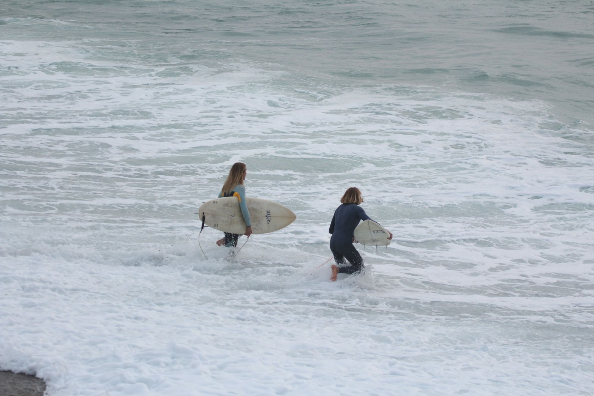 Two surfers paddle on choppy waves, each holding a surfboard in the ocean.