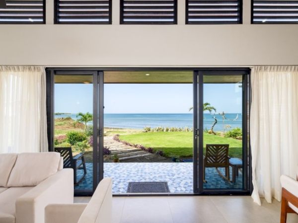 A bright living room with sofas opening onto a patio garden, where the ocean and sky are visible through large sliding doors.