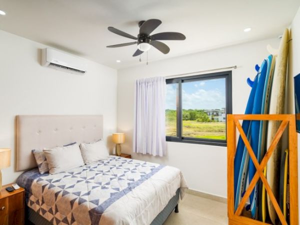 A bright bedroom with a bed, ceiling fan, large window view, and a rack holding blue surfboards.