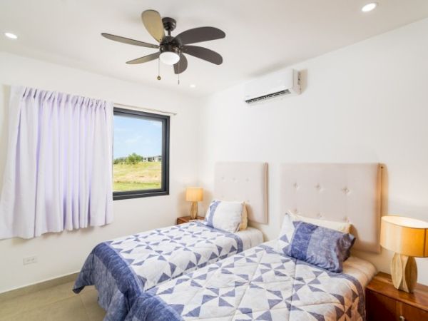 A bright bedroom with two beds, a ceiling fan, air conditioner, lamps, and blue patterned bedding by a window ending the sentence.