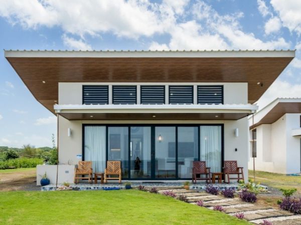 A modern white house with a large glass front, outdoor seating, and manicured garden under a blue sky.