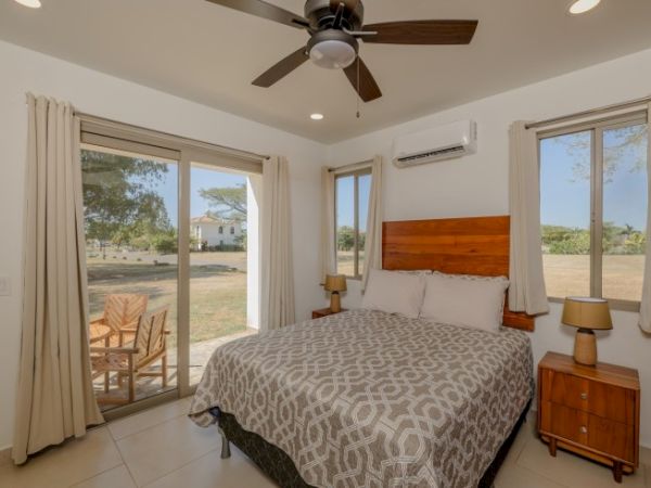 A bright bedroom with a large bed, wood headboard, curtains, ceiling fan, and two wide windows overlooking the yard.