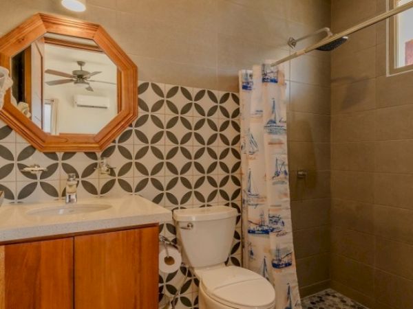 A modern bathroom with a patterned tile wall, wooden vanity and mirror, a toilet, and a shower with a blue curtain.