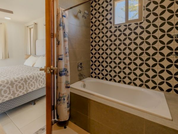 A modern bathroom with a white bathtub, patterned tile wall, window, and a glimpse of a bedroom with a bed and curtains.