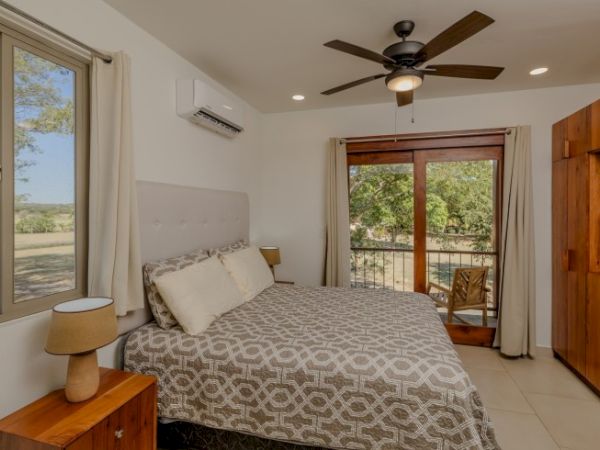 A bright bedroom with a ceiling fan, air conditioner, windows, a bed, nightstands, and a wooden door leading to a balcony.
