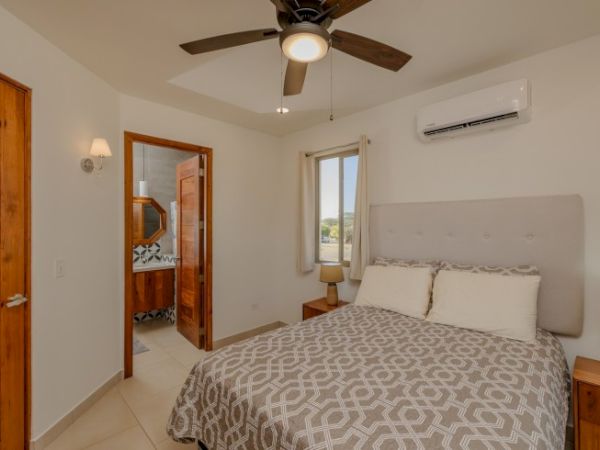 A bright bedroom with a bed, patterned bedding, window, ceiling fan, air conditioner, and wooden dresser.