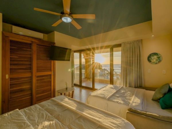 A bright hotel bedroom with two beds, a ceiling fan, and large sliding doors opening to a balcony.