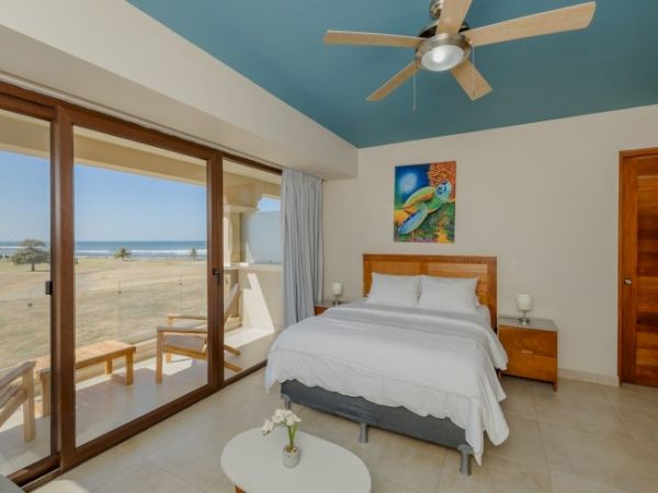 A bright beachfront bedroom with a white bed, ceiling fan, and large windows showing sandy ocean views.