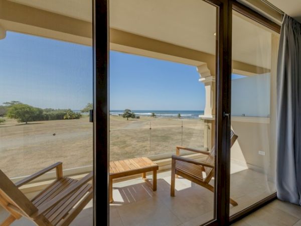 An open terrace with wooden chairs looks out to a sandy lawn and the ocean under a clear blue sky.