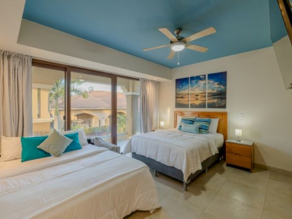 A bright bedroom with two beds, a ceiling fan, sliding glass doors to a patio, and beach-themed wall art.