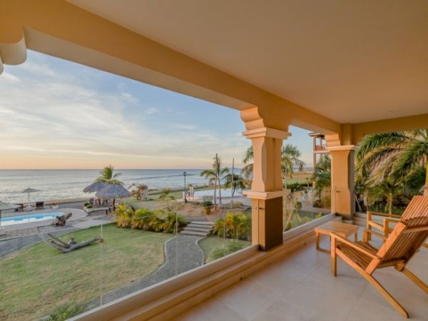 Oceanfront hotel balcony with wooden chairs, palm trees, a pool, and a sunset over the sea.