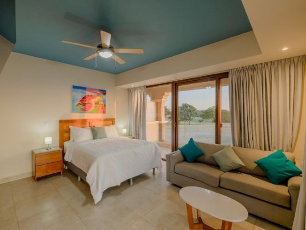 A modern bedroom with a bed, ceiling fan, and a living-area sofa, plus large glass doors opening to a view.