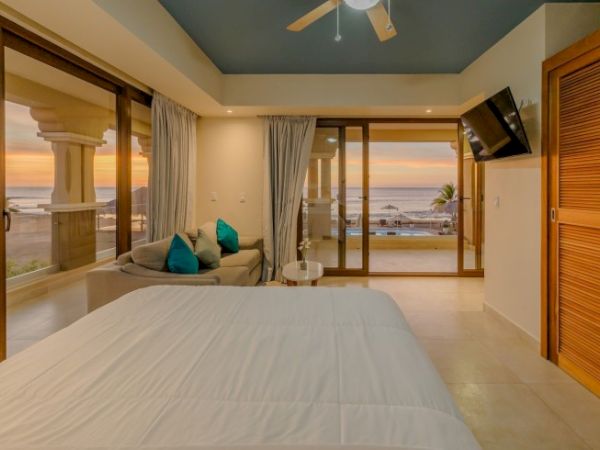 A luxurious ocean-view bedroom with a king bed, lounge seating, large sliding doors, and a ceiling fan with sunset light.