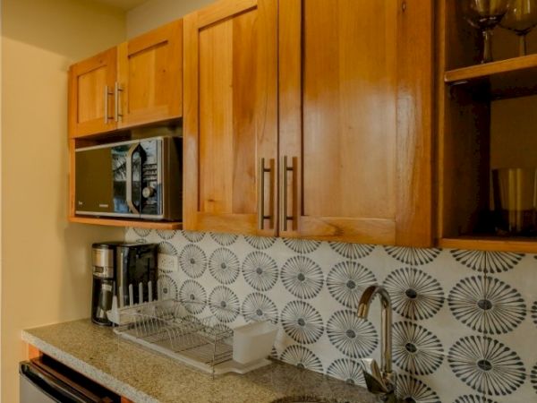A small kitchen with wooden cabinets, a microwave, dish rack, and a black-and-white circular tile backsplash.