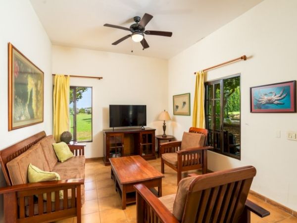 Sunny living room with wooden furniture, TV stand, ceiling fan, and framed art beside large yellow-curtained windows.