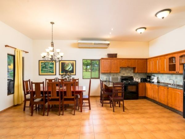 A bright dining and kitchen area with wooden chairs, pendant lights, framed ocean art, and built-in cabinets and appliances.