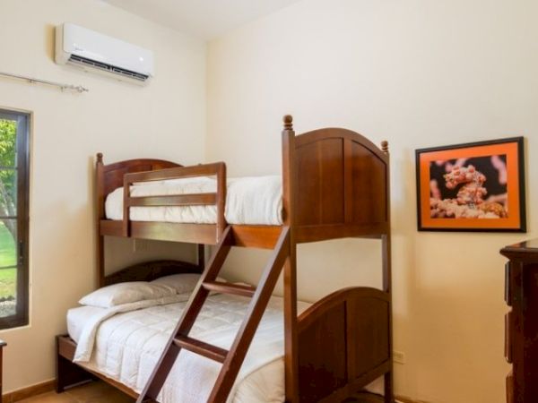 A small bedroom with a wooden bunk bed, air conditioner, window, and framed floral artwork on the wall.
