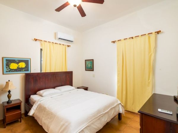 A simple bedroom with a wooden bed, yellow curtains, ceiling fan, wall AC, and bedside tables.