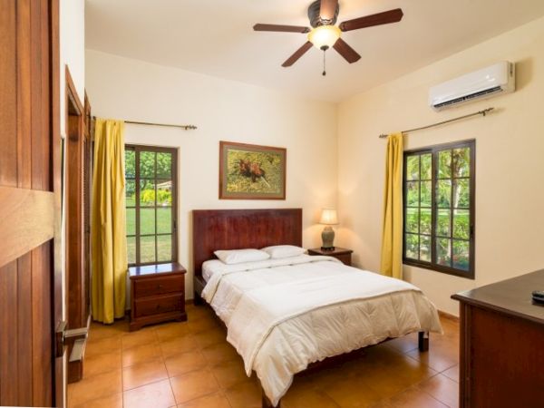 Bright bedroom with a double bed, wooden furniture, yellow curtains, wall art, and a ceiling fan.