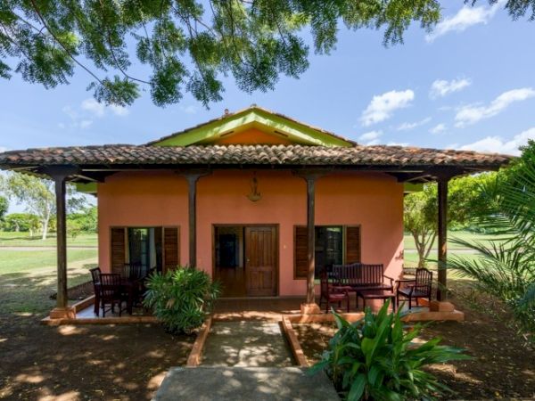 A single-story house with a tiled roof, shaded front porch, and tropical gardens with trees and shrubs around it.